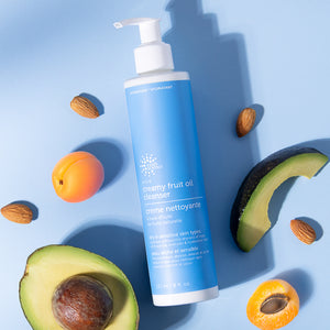 Blue bottle of creamy fruit oil cleanser surrounded by avocado, apricot, and almonds on a blue background