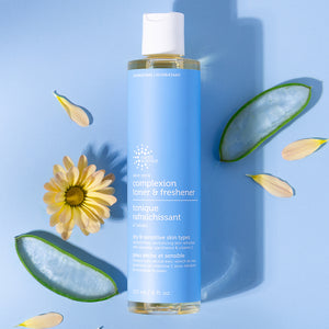 Blue bottle of 'complexion toner & freshener' with aloe vera leaves and a flower on a blue background