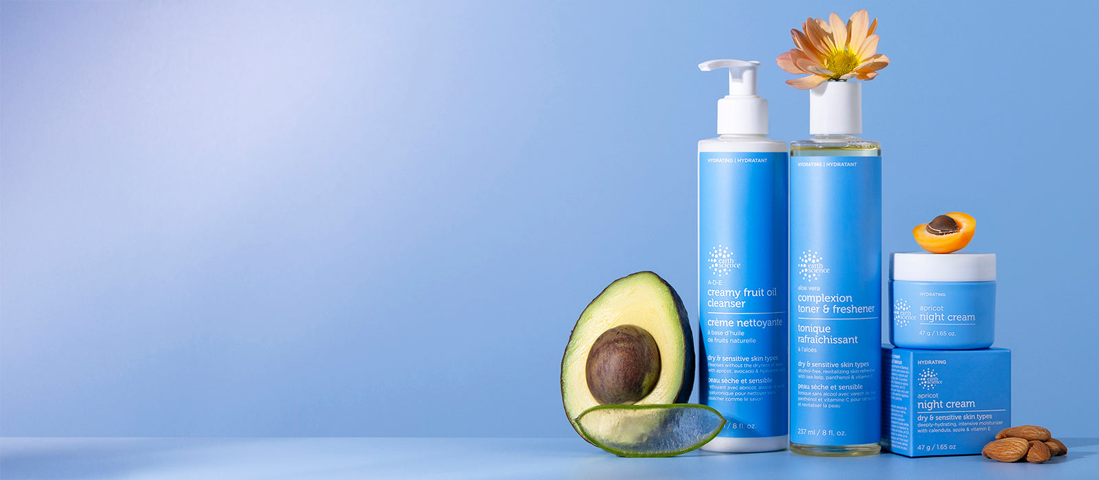 A set of blue hydrating skincare products including a pump bottle, a jar, and a small box, styled with avocado halves, almonds, and a flower on a light blue background.